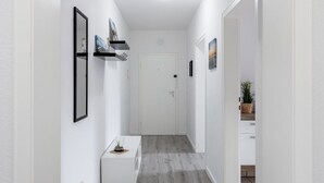 Um corredor com uma porta branca no final, uma pequena mesa branca com uma planta e um espelho na parede à esquerda.
