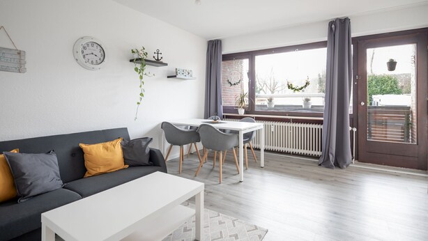 Uma sala de estar moderna com um sofá, uma mesa de jantar e cadeiras. Há um relógio branco na parede e uma janela com cortinas.