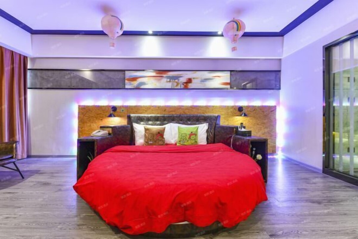 A modern bedroom with a large bed, a headboard with built-in lighting, a nightstand with a lamp, and a large window with a view.
