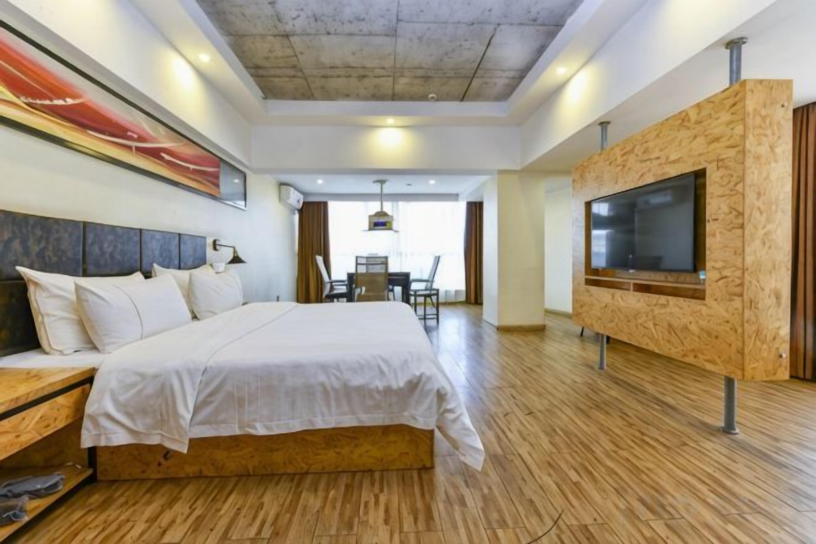 A hotel room with a large bed, a wooden TV stand, and a dining area in the background.