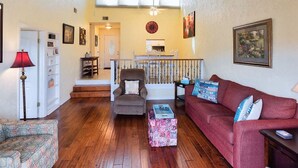 Ein Wohnzimmer mit Holzdielen, einem roten Sofa, einem gemusterten Sessel, einem Couchtisch und einer Treppe, die in einen oberen Bereich führt.