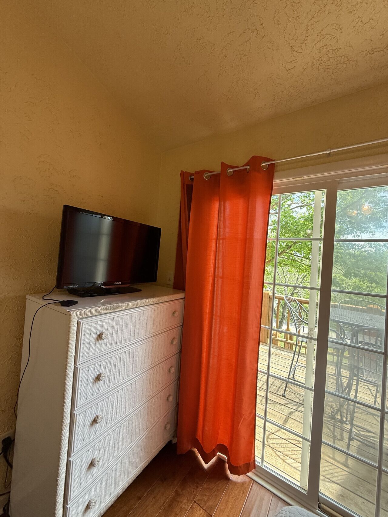 A room with a television on a dresser, a window with orange curtains, and a view of an outdoor deck.