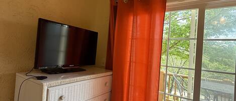 A room with a television on a dresser, a window with orange curtains, and a view of an outdoor deck.