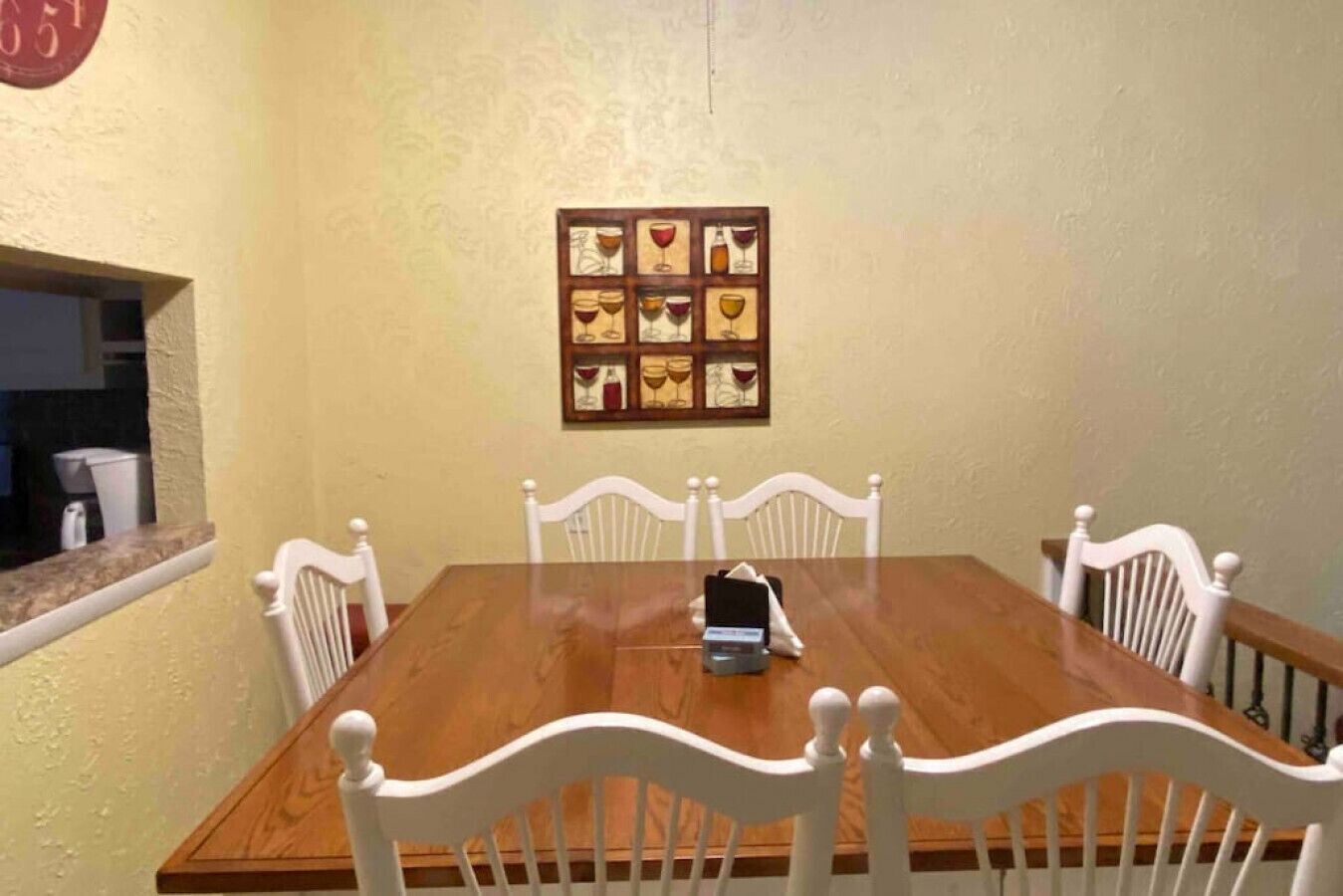 A dining room with a wooden table and white chairs, a framed picture of wine glasses on the wall, and a clock above the door.