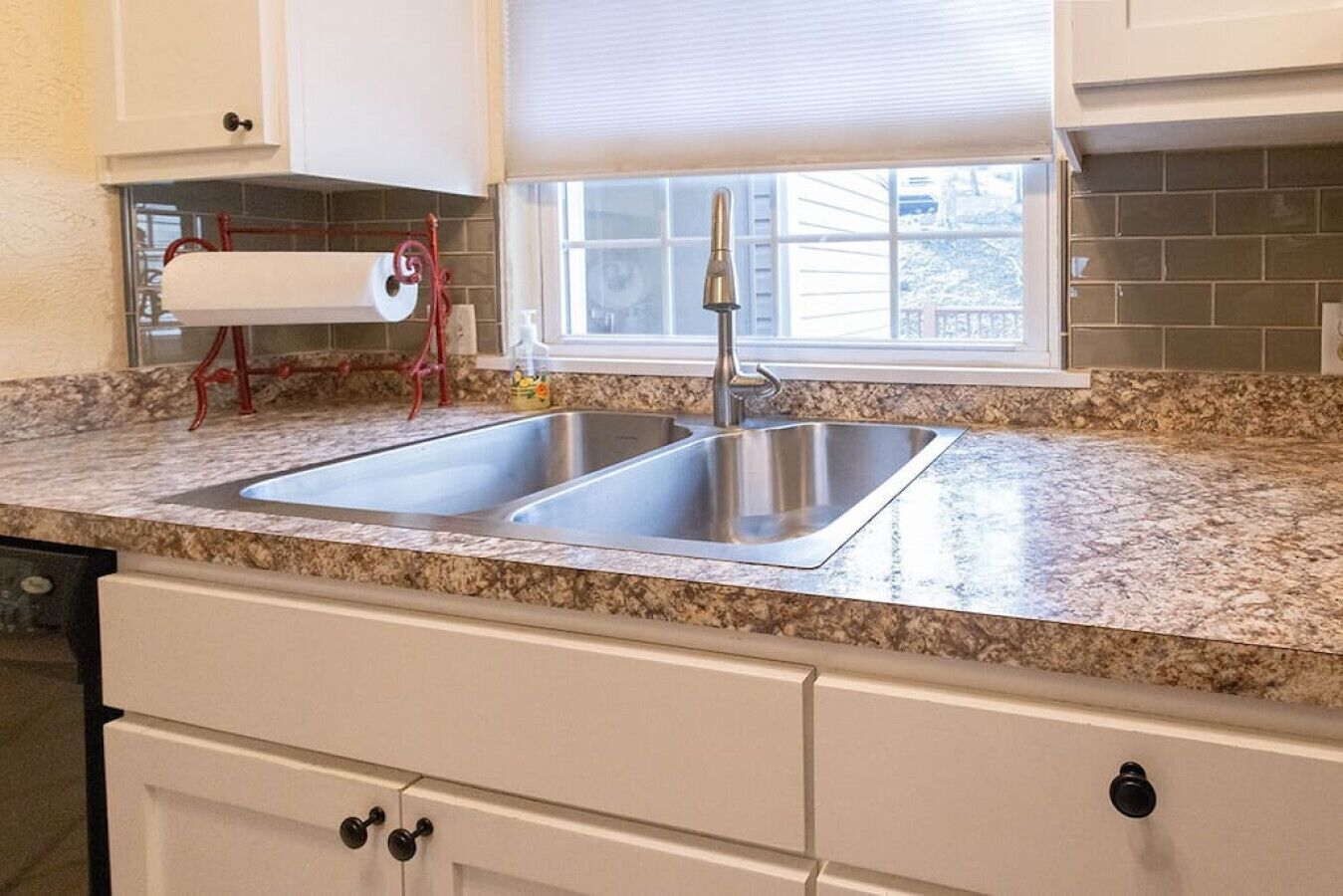 A kitchen with a double stainless steel sink, granite countertop, white cabinets, and a window with blinds.