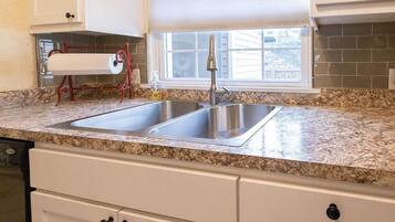A kitchen with a double stainless steel sink, granite countertop, white cabinets, and a window with blinds.