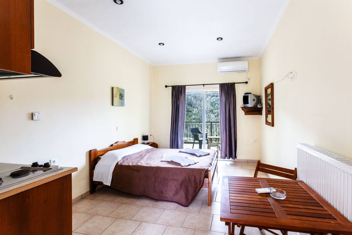 A bedroom with a bed, a wooden desk, a table, and a balcony with a chair.