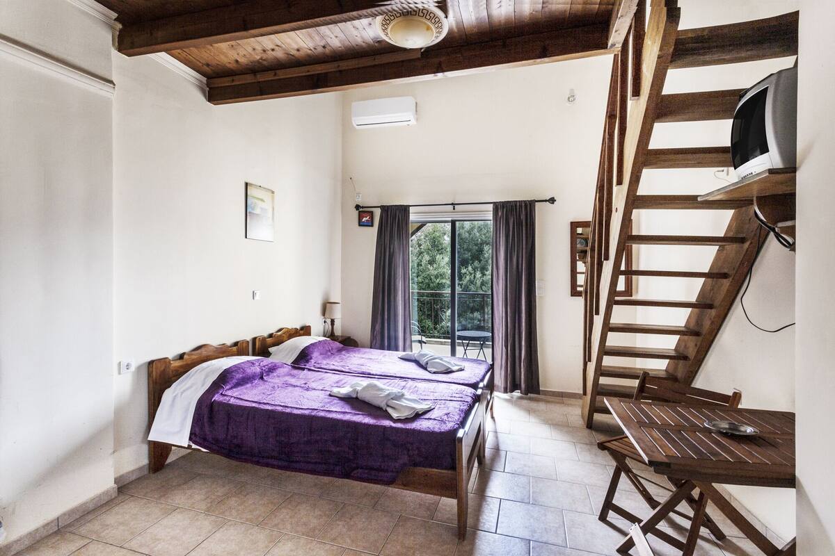A bedroom with two wooden beds, a balcony with a table, and a TV mounted on a wooden beam.