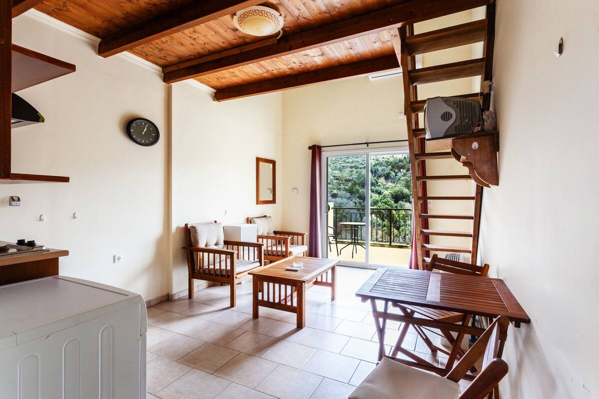 A room with a wooden ceiling, a large window with a view of greenery, a table with chairs, and a ladder against the wall.
