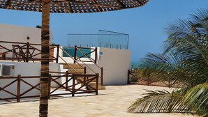 A covered outdoor area with a patterned umbrella, palm trees, and a white building with a glass enclosure. - Peixe Gordo Hotel (Icapuí)