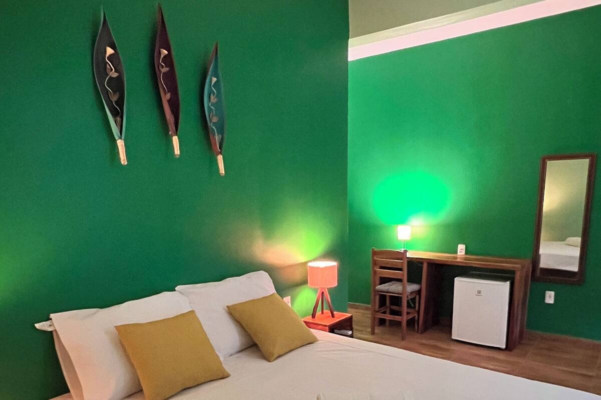 A hotel room with a large bed, green walls, a desk with a lamp, and a small refrigerator.