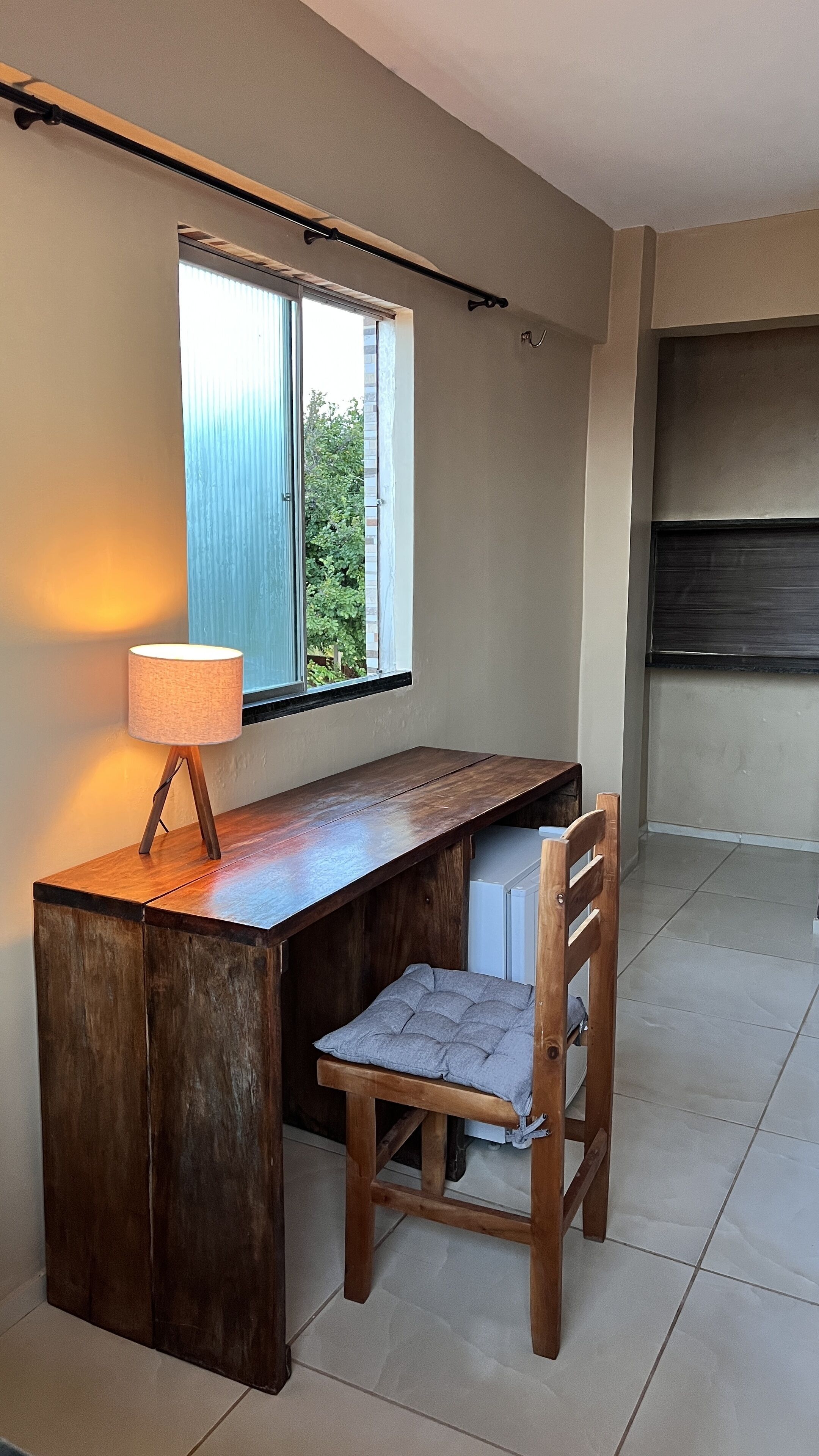 Uma mesa de madeira com uma cadeira, uma luminária e uma janela com vista para o verde.