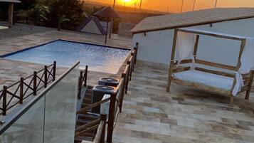 Uma área de piscina no terraço com guarda-corpo de vidro, um banco de madeira e vista para turbinas eólicas ao fundo durante o pôr do sol.