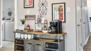 Un coin cuisine avec un espace dédié au café, un panneau d’accueil et des œuvres d’art encadrées accrochées au mur.