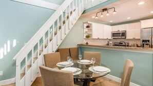 A dining area with a round glass table set for four, surrounded by beige chairs. A kitchen with white cabinets and stainless steel appliances is visible in the background. - Headline (Sanibel)