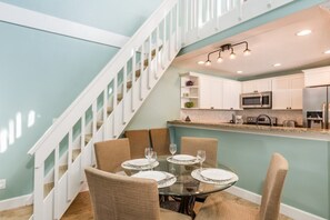 A dining area with a round glass table set for four, surrounded by beige chairs. A kitchen with white cabinets and stainless steel appliances is visible in the background.