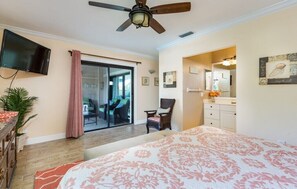 A bedroom with a bed, a ceiling fan, a TV, a sliding glass door, a chair, a dresser, and a plant.