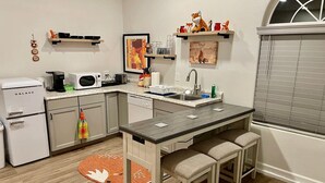 A compact kitchen with a central island, a refrigerator, and a microwave. There are two stools at the island, and a window with blinds to the right.