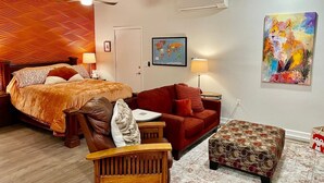 A bedroom with a bed, a red sofa, a wooden chair, and a small ottoman. There is a world map on the wall and a painting of a fox.