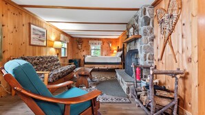 A cozy cabin interior with a stone fireplace, wooden walls, a sofa, a rocking chair, and a log bed. - Balsam Breeze - cozy cottage on Mooselookmegunic! (Rangeley)