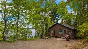 A cabin with a gravel driveway, surrounded by trees and a body of water. - Balsam Breeze - cozy cottage on Mooselookmegunic! (Rangeley)
