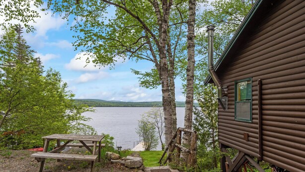 A wooden cabin with a view of a lake, a picnic table, and a wooden deck. - Balsam Breeze - cozy cottage on Mooselookmegunic! (Rangeley)