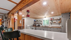 A kitchen with a wooden ceiling, white countertops, and grey cabinets. There is a stove, a sink, and a coffee maker. - Balsam Breeze - cozy cottage on Mooselookmegunic! (Rangeley)