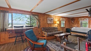 A wooden cabin interior with a sofa, chairs, a bed, and a view of the outdoors. - Balsam Breeze - cozy cottage on Mooselookmegunic! (Rangeley)