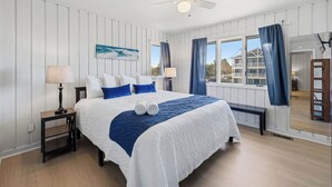 A bedroom with a bed, a ceiling fan, a window with curtains, and a view of a building outside. - Cair Paravel | 680 ft to the beach | (Virginia Beach)