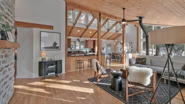 A spacious living area with wooden flooring, a large sofa, a fireplace, and a view of the kitchen and dining area through large windows. - Durango Mountain House with Hot Tub and Views - great location (Durango)