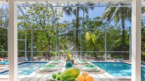A poolside dining area with a table set for two, surrounded by a glass enclosure with a view of palm trees and a pool. - ***NEW*** Beautiful Cottage Canal Front&Boat Dock (Sanibel)