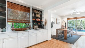 A modern kitchen with white cabinetry, a dining area with a wooden table and chairs, and a large window with a view of the outdoors. - ***NEW*** Beautiful Cottage Canal Front&Boat Dock (Sanibel)