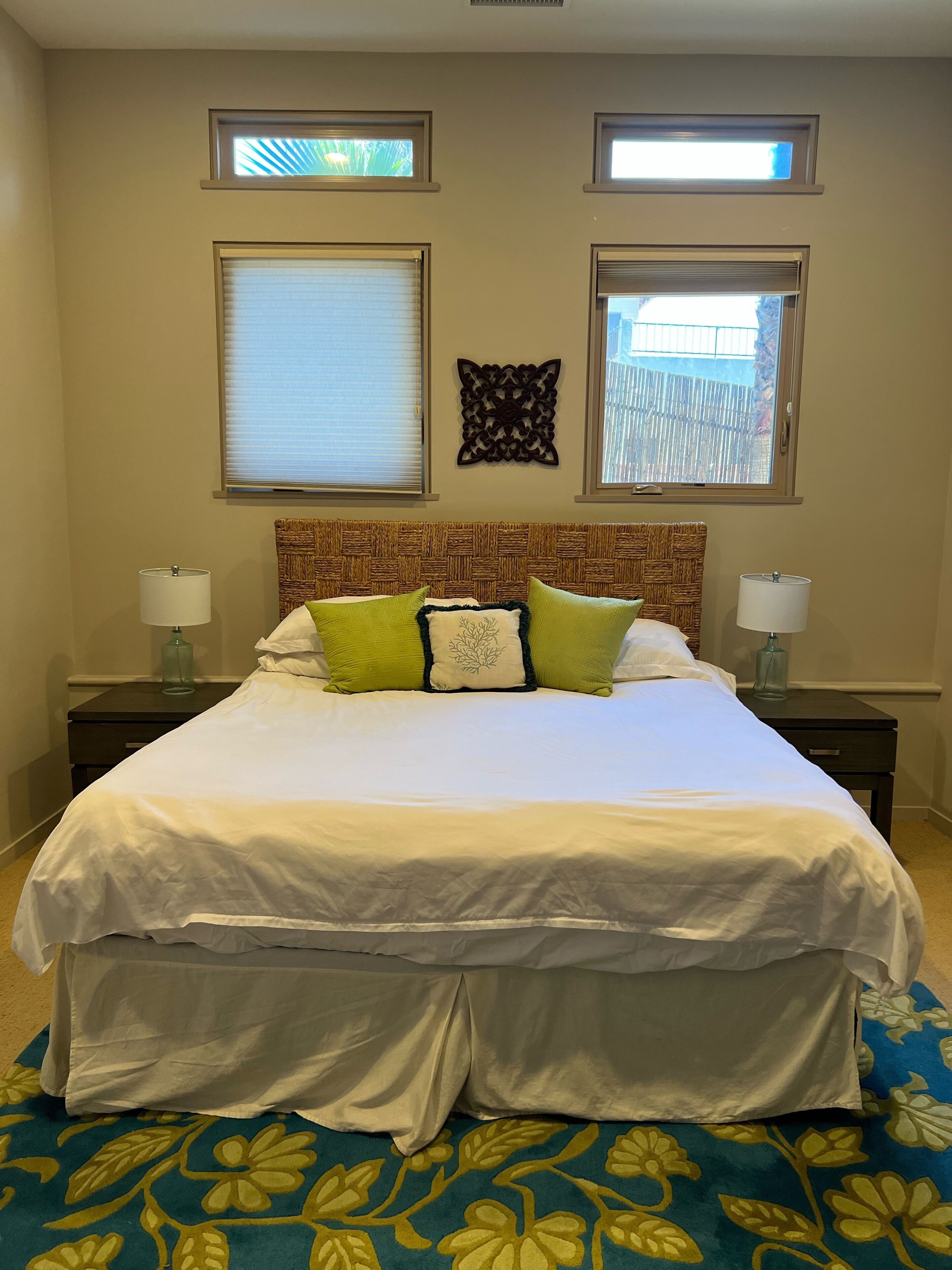 A bedroom with a large bed, two bedside tables, a decorative headboard, and two windows with blinds.