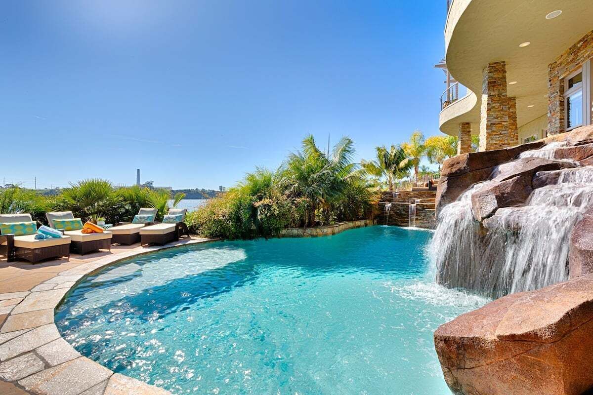A clear blue pool with a waterfall feature, surrounded by lounge chairs and palm trees.