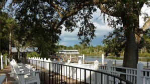 A waterfront patio with white rocking chairs and tables, a white fence, and a building in the background. - Redfish Lodge- Steinhatchee Landing Resort Sleep 5 (Steinhatchee)