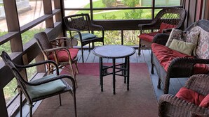 A covered porch with wicker furniture, a small round table, and a view of a red house and greenery. - Redfish Lodge- Steinhatchee Landing Resort Sleep 5 (Steinhatchee)