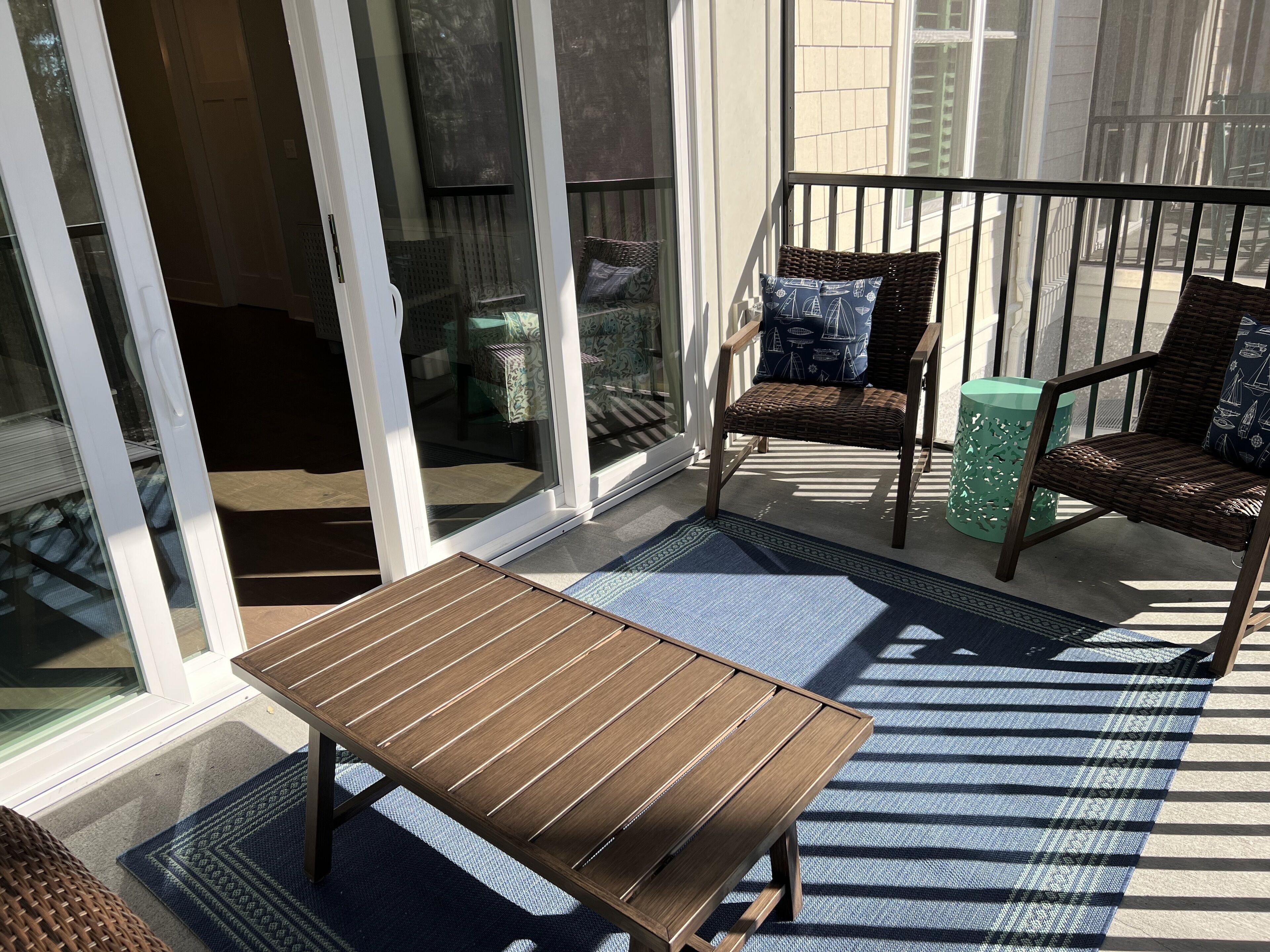 A sunlit patio with a wooden table, two wicker chairs, and a green decorative stool.