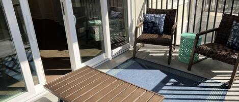 A sunlit patio with a wooden table, two wicker chairs, and a green decorative stool.