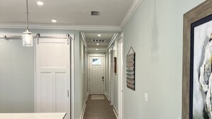 Un couloir avec un coin cuisine sur la gauche, comprenant un comptoir blanc et des tabourets de bar. Les murs sont d’un bleu clair, et un tableau abstrait encadré est accroché au mur de droite.