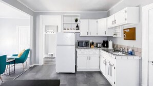 A modern kitchen with white cabinetry, a refrigerator, a microwave, and a coffee maker. There is a dining area with a teal chair and a small table. - Blocks from River & DT Missoula! University apt now offering mid-term rentals! (Missoula)