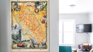 A room with a large map on the wall, a wooden bench, a dining table, teal chairs, and a flat-screen TV mounted on the wall. - Blocks from River & DT Missoula! University apt now offering mid-term rentals! (Missoula)