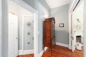 A hallway with a wooden floor, a white door, a wall with floral wallpaper, a tall wooden cabinet, and a fireplace with a white mantel.