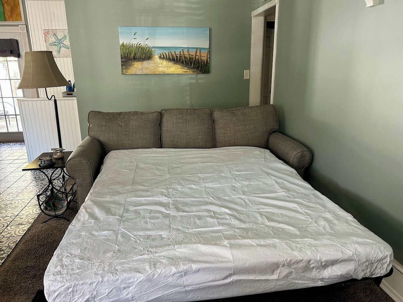 A bedroom with a large bed, a painting of a beach scene, and a lamp on a side table.