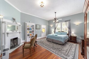 A bedroom with a fireplace, a bookshelf, a bed with a blue bedspread, and a large window with curtains. - Private historic district apt with pool-suite A (Ocala)