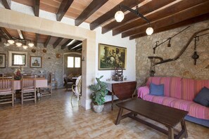 Un interior rústico con vigas de madera a la vista, paredes de piedra, una zona de comedor con muebles de madera y un rincón acogedor con un sofá a rayas.