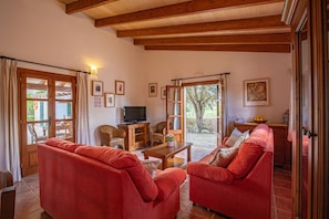 A living room with two red sofas, a wooden coffee table, and a television on a wooden stand. There are framed pictures on the wall, a window with curtains, and a door leading outside.