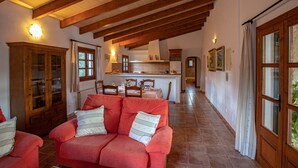 A spacious living area with a red sofa, wooden beams on the ceiling, and a kitchen in the background. - Lovely villa in Pollensa countryside (Pollensa / Pollença)