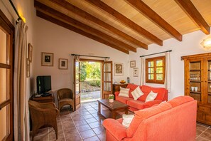 Ein gemütliches Wohnzimmer mit einem roten Sofa, einem Holztisch und Korbstühlen. Ein Fernseher steht auf einem kleinen Tisch, und ein Holzschrank beherbergt Gläser.