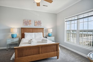 A bedroom with a wooden bed, two bedside tables with lamps, a large window with a view, and two wall art pieces.
