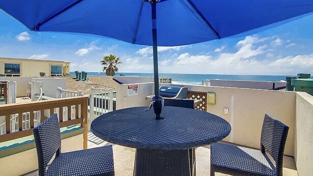 A balcony with a round table and chairs, a blue umbrella, and a view of the ocean.